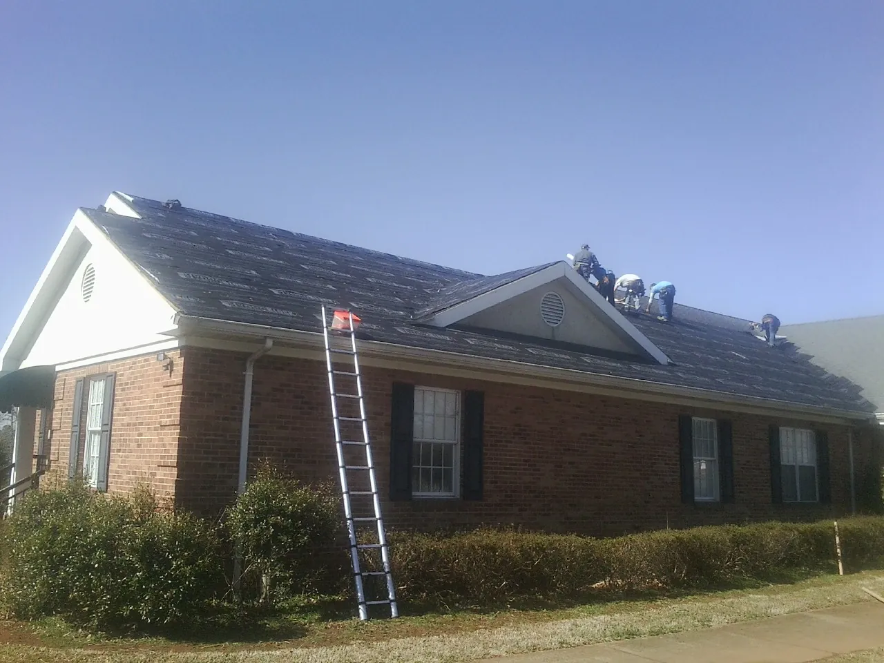 Crew tearing shingles off a brick ranch roof with a ladder set — Metal Roof Repair work by our crew in Newfane