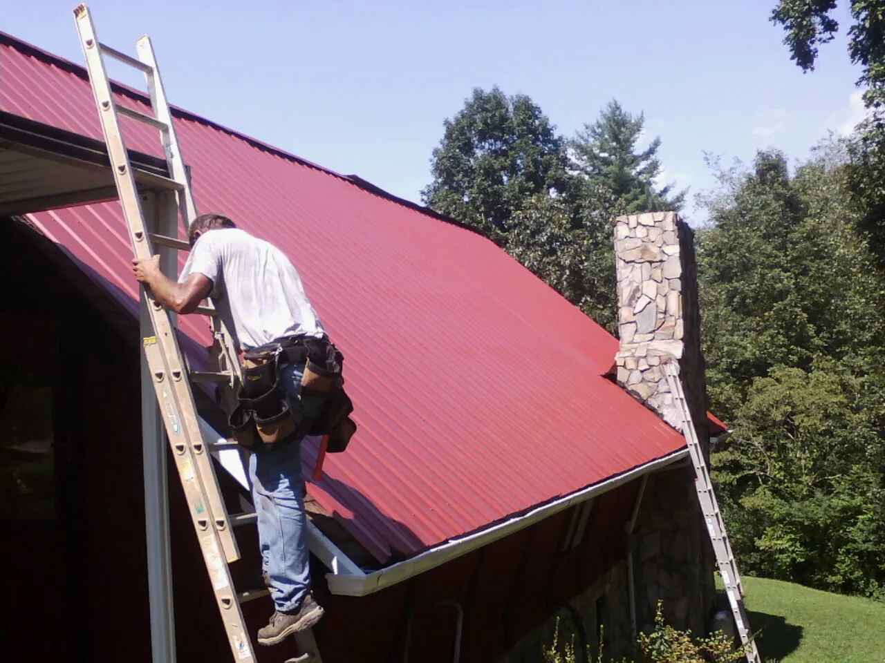 Roofer on a ladder beside a red metal roof and stone chimney — Soffit & Fascia Repair by Roofing Brothers in Newfane